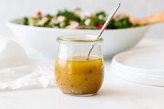 a glass jar filled with dressing next to a bowl of salad on a white table