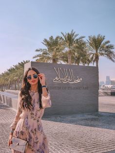 a woman standing in front of a sign with palm trees behind her and wearing sunglasses