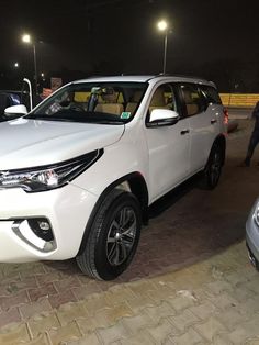 a white suv is parked on the side of the road at night with people walking by