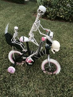 a skeleton on a small bike in the grass next to a ball and some scissors