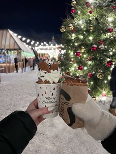 someone is holding up a cup of ice cream in front of a christmas tree at night