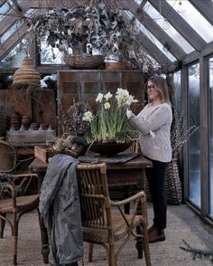 a woman standing next to a table with flowers on it in a room filled with plants