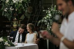 two people sitting at a table with flowers and greenery