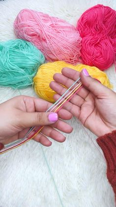 two hands holding yarn and knitting needles in front of balls of yarn on a white blanket