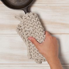 a hand holding a knitted dish cloth next to a frying pan on a wooden table