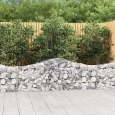 a stone wall in front of a wooden fence with trees and shrubbery behind it