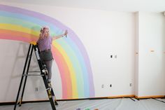 a woman standing on a ladder painting a rainbow wall