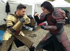 two men dressed in armor standing next to each other on gravel ground with tents behind them