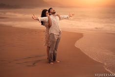 a man and woman standing on top of a beach next to the ocean at sunset