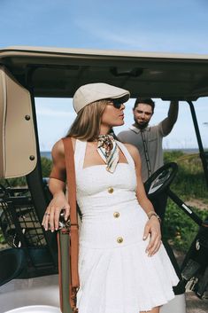 a woman in a white dress and hat standing next to a golf cart with a driver