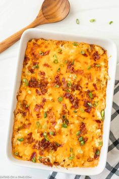 a casserole dish with cheese, bacon and green onions next to a wooden spoon