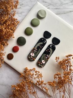 three black and green earrings sitting on top of a white board next to dried flowers