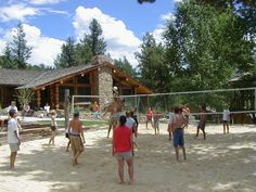 several people are playing volleyball in the sand near a log cabin and trees, while others watch