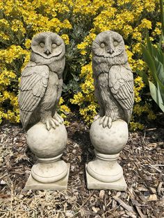 two statues of owls sitting on top of each other in front of flowers and bushes