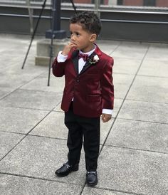 a young boy dressed in a red suit and bow tie standing on the sidewalk with his hand to his mouth