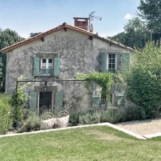 an old stone house with green shutters on the front and side windows, surrounded by greenery