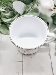 a white cup sitting on top of a wooden table next to green leaves and cotton