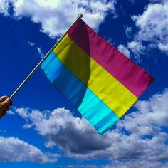 a hand holding a rainbow colored flag in the air with clouds behind it on a sunny day