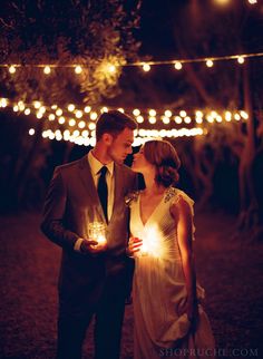 a man and woman standing next to each other holding candles in their hands at night