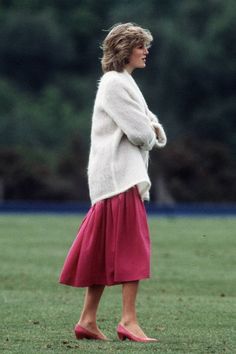 a woman in a red skirt and white sweater is standing on the grass with her hands behind her back