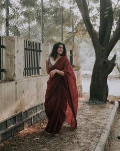 a woman in a red sari is standing near a tree and fence with her hands on her hips