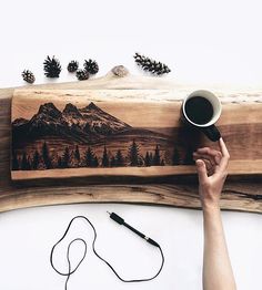 a person holding a coffee cup next to a piece of wood with mountains and pine cones on it