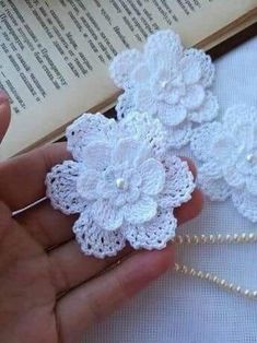 white crocheted flowers are being held in front of an open book with pearls