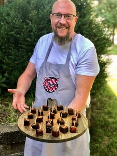 a man in an apron is holding a tray with small desserts on it and pointing at the camera