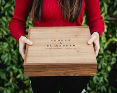 a woman holding a wooden box with the name and date engraved on it, in front of some bushes