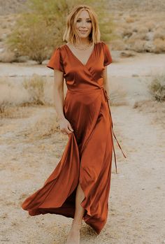 a woman standing in the desert wearing an orange wrap dress and tan sandals with her legs crossed