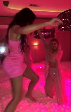 two women are dancing in the middle of a room with pink lights and white carpet