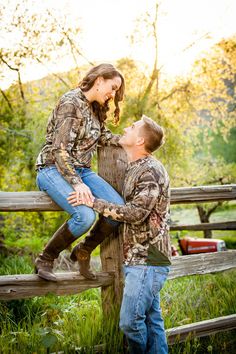 a man and woman sitting on top of a wooden bench in the grass with trees behind them