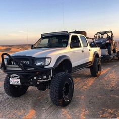 a white truck parked in the desert next to two other trucks and one is pulling a buggy