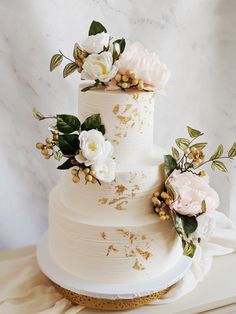 a three tiered white wedding cake with flowers on top and gold leaf decoration around the edges