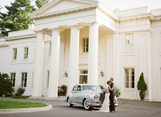 a bride and groom standing next to an old car in front of a large white building