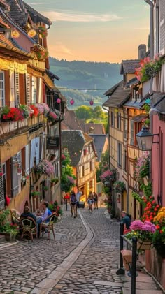 a cobblestone street with people sitting at tables and flowers on the balconies