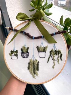 a person holding up a cross - stitch embroidery project with potted plants hanging from it's sides