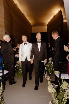 two men in tuxedos are walking down the aisle with other people behind them