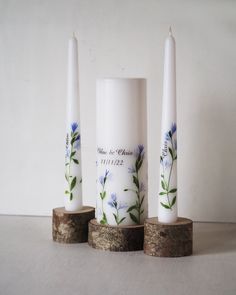 three white candles with blue flowers on them are sitting on wood slices in front of a wall