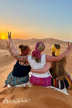 three women sitting on top of a sand dune in the middle of the desert at sunset