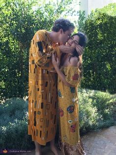 two people are hugging each other in front of some bushes and trees, one is wearing a yellow dress