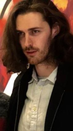 a man with long hair wearing a white shirt and black blazer standing in front of a painting