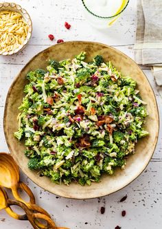 a bowl filled with broccoli salad next to two wooden spoons on a white table