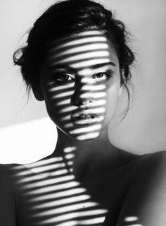 a woman standing in front of a window with the shadow of blinds on her face