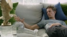 a man laying on a couch with an owl statue next to him and a glass bottle in the foreground