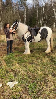 a woman standing next to a black and white horse in a field with trees behind her