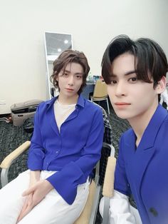 two young men sitting next to each other in an office setting, one wearing a blue shirt and the other white pants