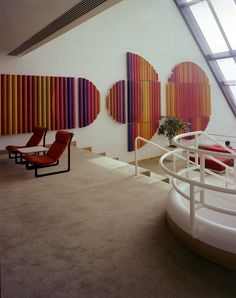 an empty room with chairs and paintings on the wall behind them, in front of a spiral staircase