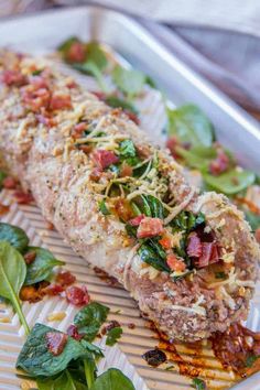 a white plate topped with meat covered in sauce and veggies next to spinach leaves