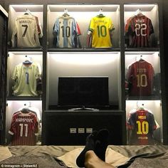 a person's feet resting on a bed in front of a display case filled with soccer jerseys